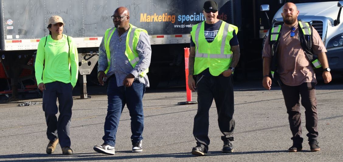 Four men wearing safety vests and work clothing walk together across a trucking yard with trailers behind them.