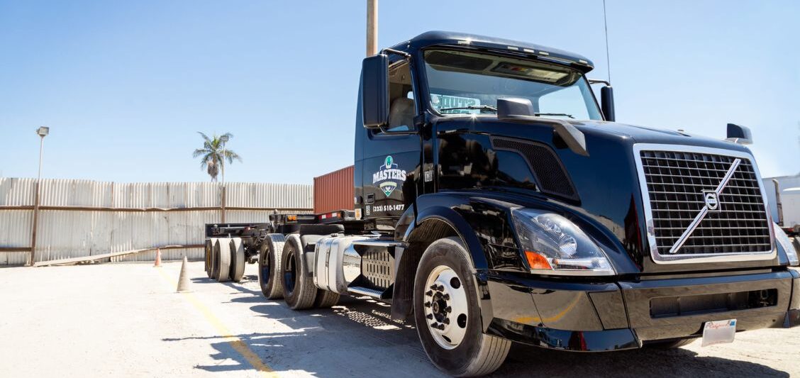 A black semi-truck sits in an outdoor lot on a sunny day, with cones, fencing, and a palm tree visible in the background.