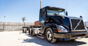 A black semi-truck sits in an outdoor lot on a sunny day, with cones, fencing, and a palm tree visible in the background.
