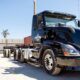 A black semi-truck sits in an outdoor lot on a sunny day, with cones, fencing, and a palm tree visible in the background.