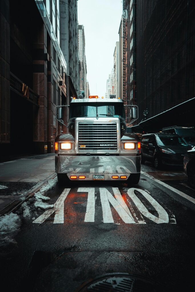 A large semi-truck is parked on a wet city street between tall buildings, with headlights on and snow melting along the road.