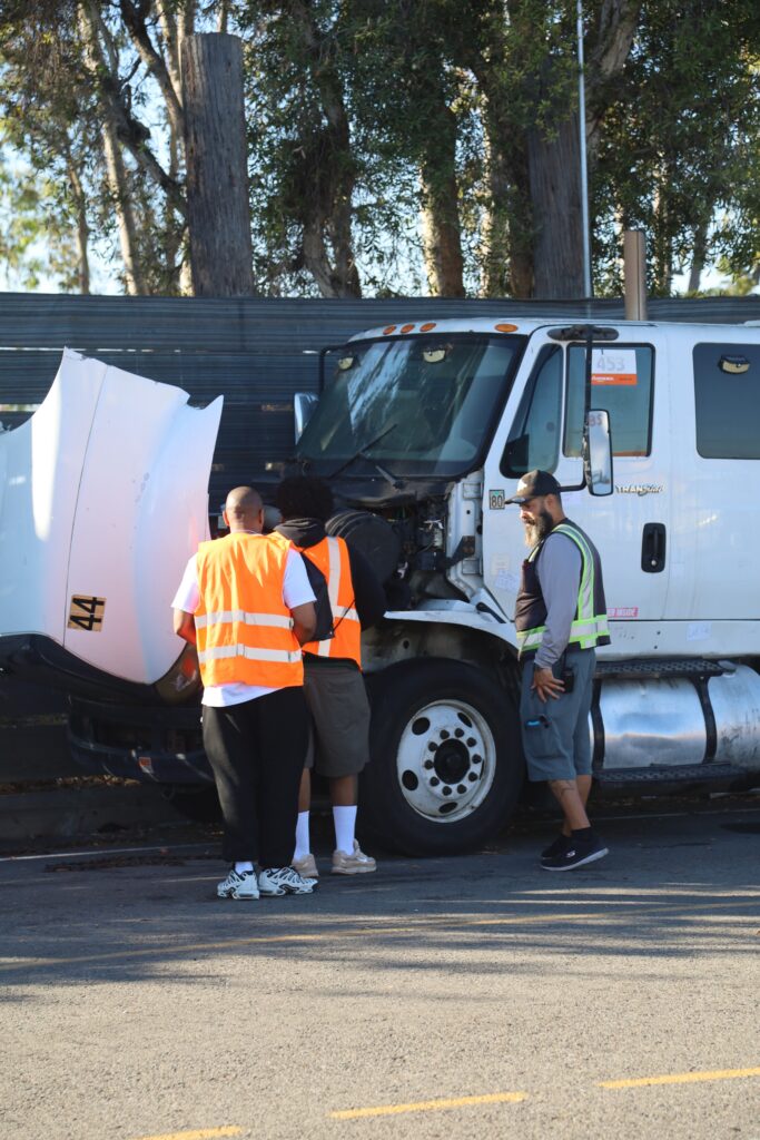 Several trainees in safety vests gather around the front of a white semi truck with the hood open while reviewing the engine area.