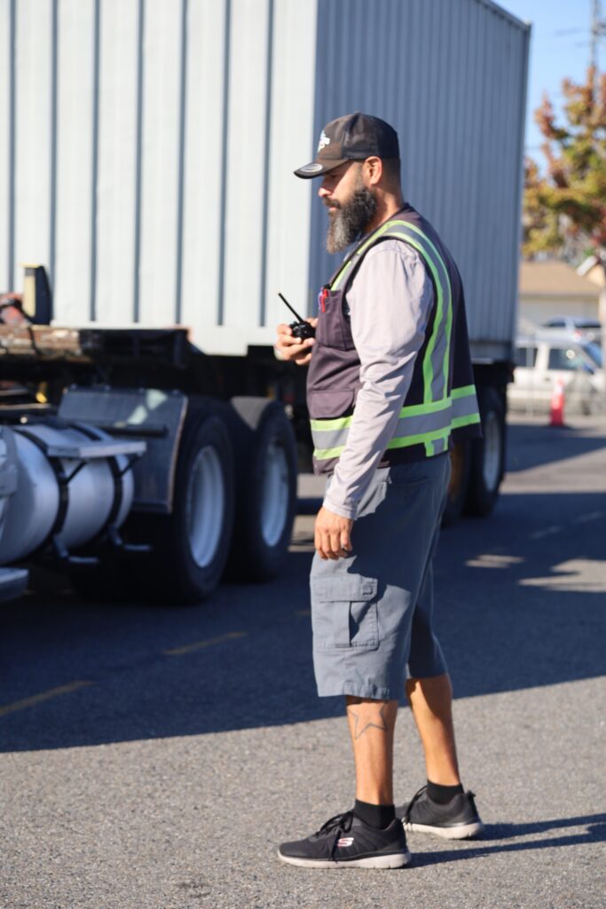 A truck driving instructor wearing a reflective vest stands beside a semi truck while holding a radio and observing a training exercise.