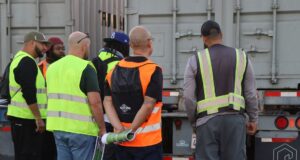 A group of truck driving students in reflective vests stand together behind a trailer while listening to an instructor during hands-on training.