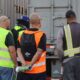 A group of truck driving students in reflective vests stand together behind a trailer while listening to an instructor during hands-on training.