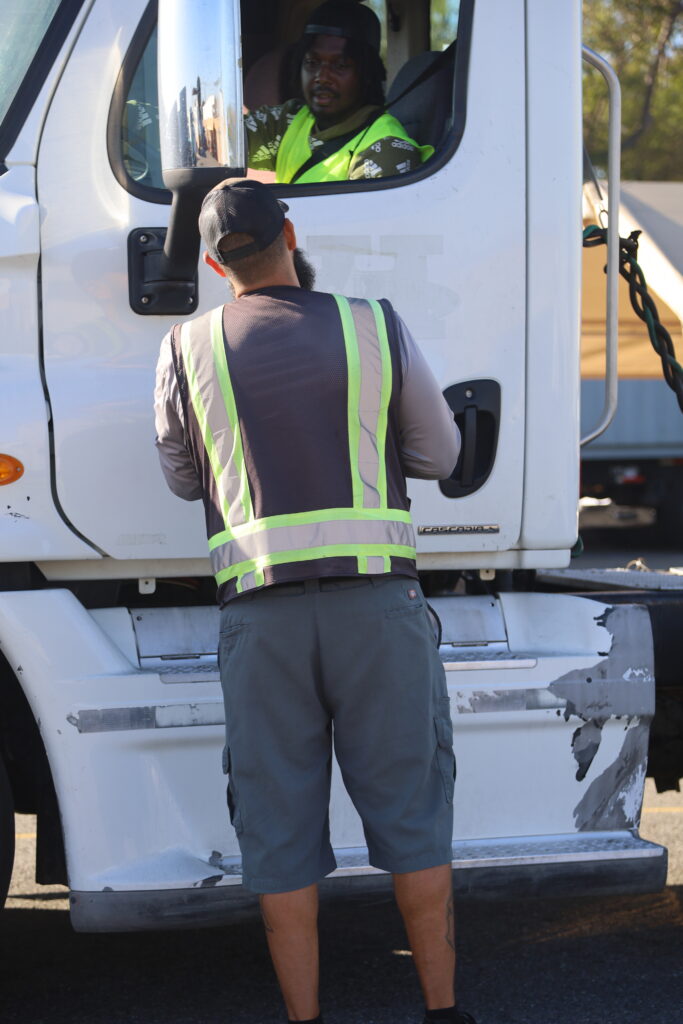 A truck driving instructor stands in a training yard wearing a reflective vest while monitoring a semi truck maneuvering nearby.
