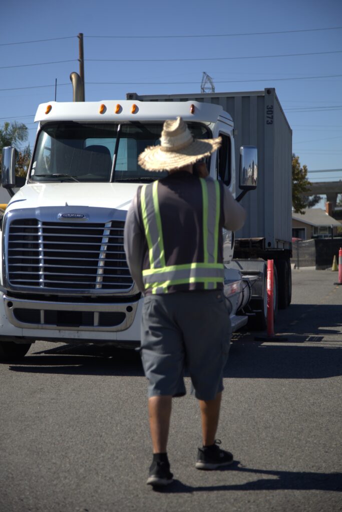 A truck driver wearing a reflective safety vest walks toward a white semi truck parked in a training lot with traffic cones nearby.