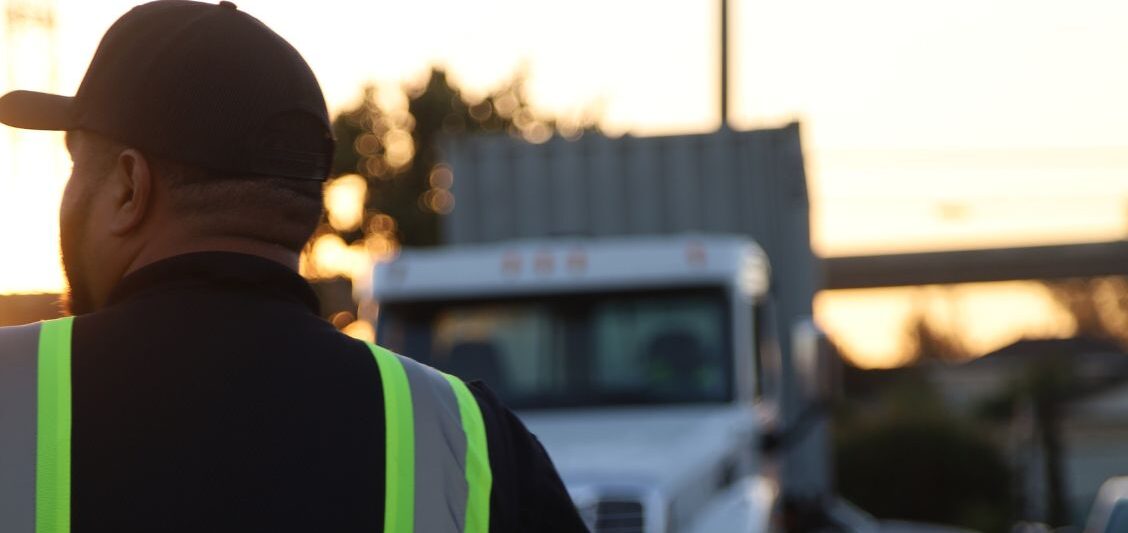 A truck driver in a reflective vest walks toward a semi truck in a training yard during daylight hours.