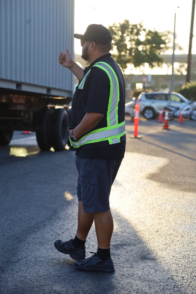 A truck driving instructor wearing a reflective vest gestures with a thumbs-up signal beside a trailer during a driving exercise.
