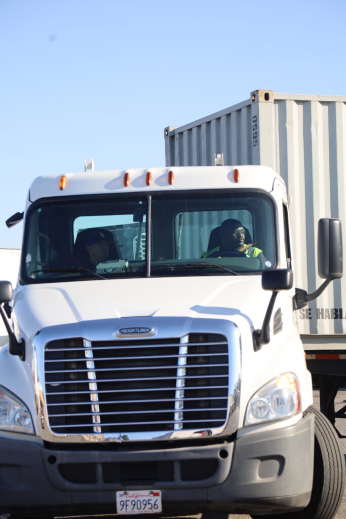 A white semi-truck with a shipping container is parked while two people sit inside the cab.