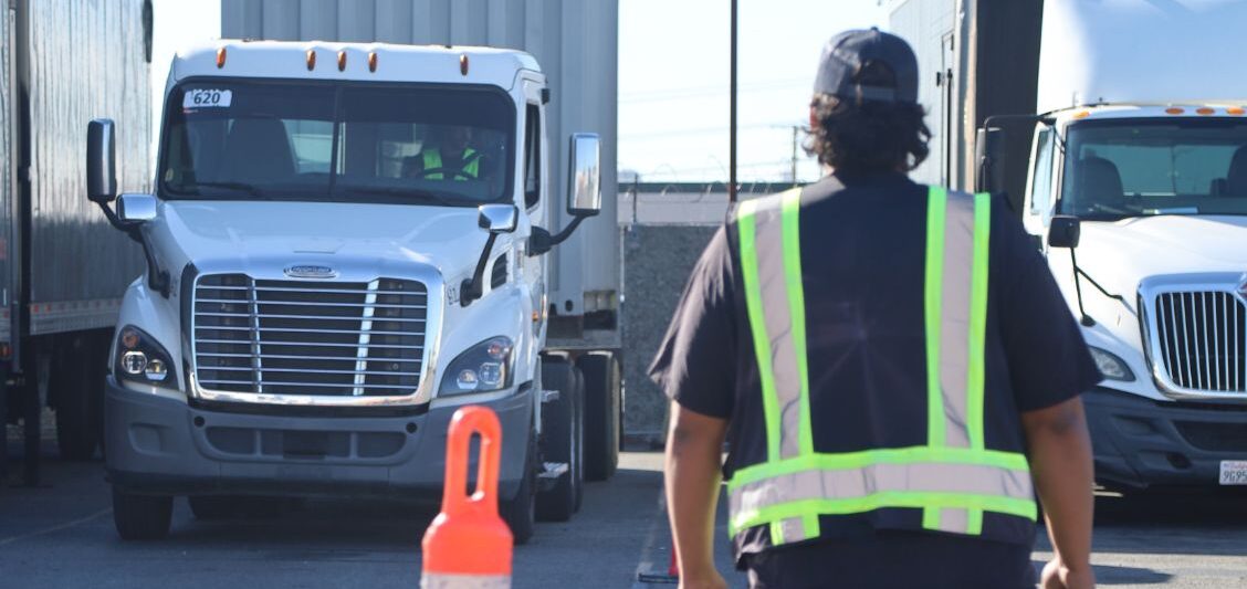 A person in a reflective safety vest walks between parked commercial trucks in a trucking yard during daylight.