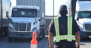 A person in a reflective safety vest walks between parked commercial trucks in a trucking yard during daylight.