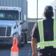 A person in a reflective safety vest walks between parked commercial trucks in a trucking yard during daylight.