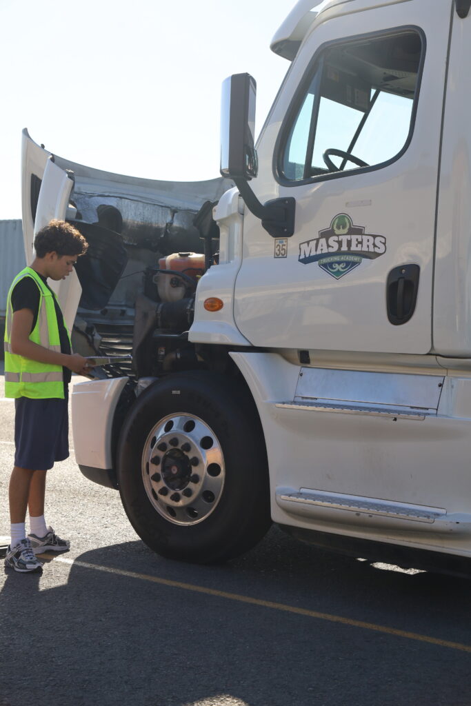A student in a reflective safety vest inspects the engine area of a white semi-truck at a trucking training yard.