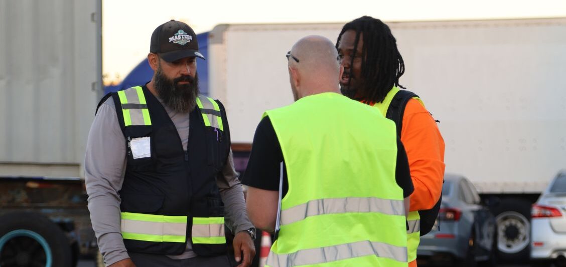Three people wearing reflective safety vests stand together in a trucking yard during a training or safety discussion.