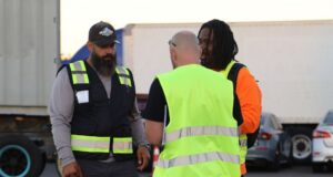 Three people wearing reflective safety vests stand together in a trucking yard during a training or safety discussion.