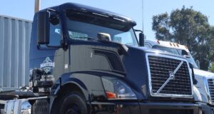 Black semi-truck cab parked in a lot beside a white trailer and another truck on a clear day.