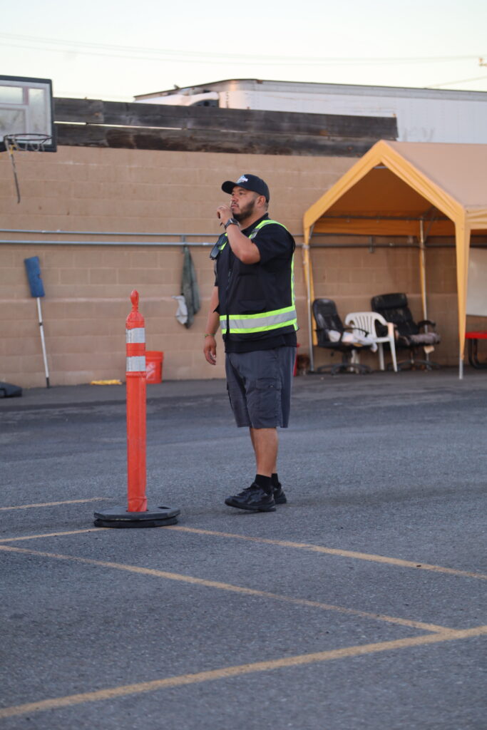 Person in a reflective safety vest standing in a training lot beside an orange traffic marker, speaking into a handheld radio.