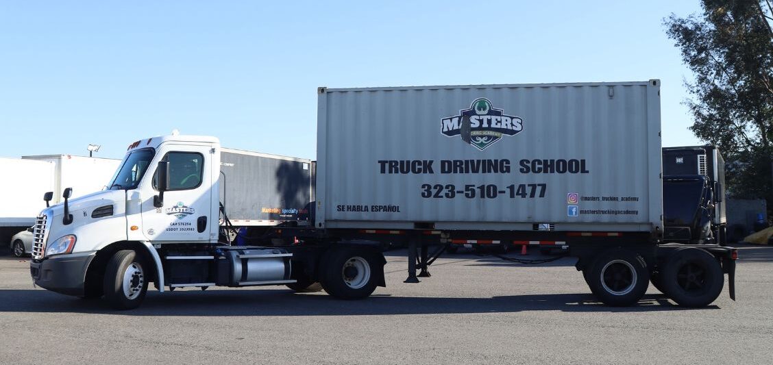 White truck and trailer parked in a training yard, with a large container displaying a trucking school name, phone number, and social media information.