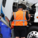 Two people inspect the engine compartment of a commercial truck with the hood raised during a maintenance check.