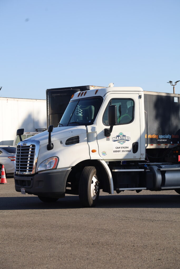 White semi-truck parked in a trucking yard with trailers and training equipment visible in the background.