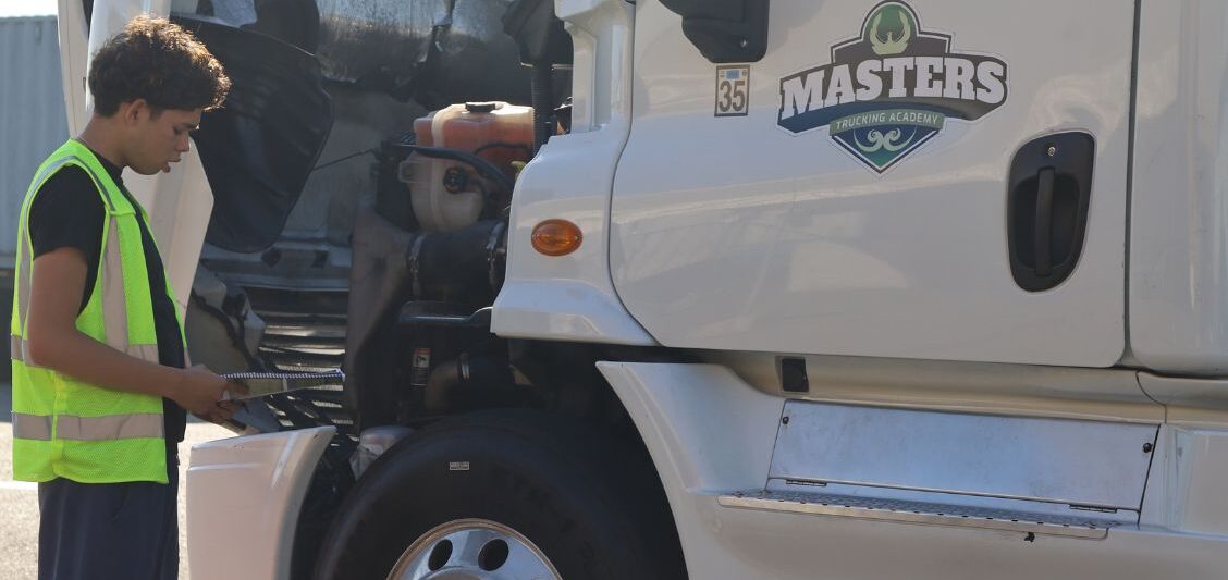 Student in a reflective safety vest reviews a checklist while inspecting the engine compartment of a white semi-truck.