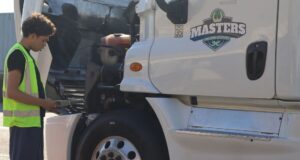 Student in a reflective safety vest reviews a checklist while inspecting the engine compartment of a white semi-truck.
