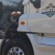 Student in a reflective safety vest reviews a checklist while inspecting the engine compartment of a white semi-truck.