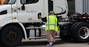 A person wearing a bright reflective safety vest stands beside the driver-side door of a white semi-truck.