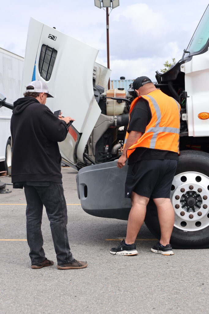 Two men stand beside a white truck with its hood open, with one wearing an orange reflective safety vest while inspecting the engine.
