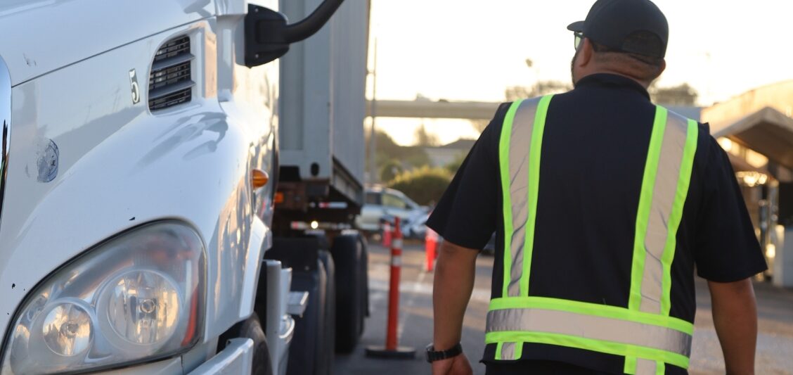 A man in a reflective safety vest walks alongside the front of a white commercial truck in an outdoor training area.