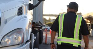 A man in a reflective safety vest walks alongside the front of a white commercial truck in an outdoor training area.