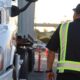 A man in a reflective safety vest walks alongside the front of a white commercial truck in an outdoor training area.
