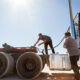 Truck driver securing equipment on a flatbed trailer at a commercial trucking facility.