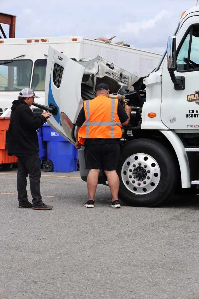 Two men inspect the open engine compartment of a white commercial truck, with one wearing a bright orange safety vest.