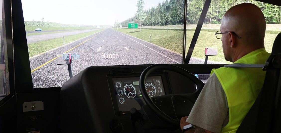 A man wearing a high-visibility vest sits at the controls of a truck driving simulator displaying a highway scene.